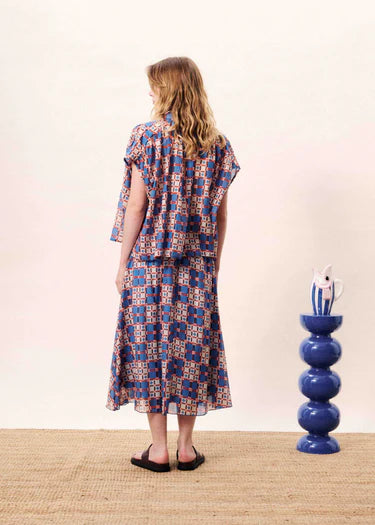 Woman wearing a patterned dress standing in front of a blue vase on a beige surface.