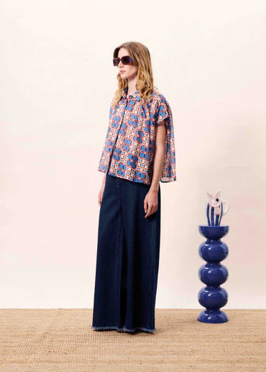 Woman wearing a patterned shirt and dark pants standing next to a decorative blue vase on a beige floor.