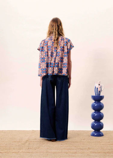 Person wearing a patterned shirt and dark pants standing in front of a white wall with a blue vase on a wooden surface.