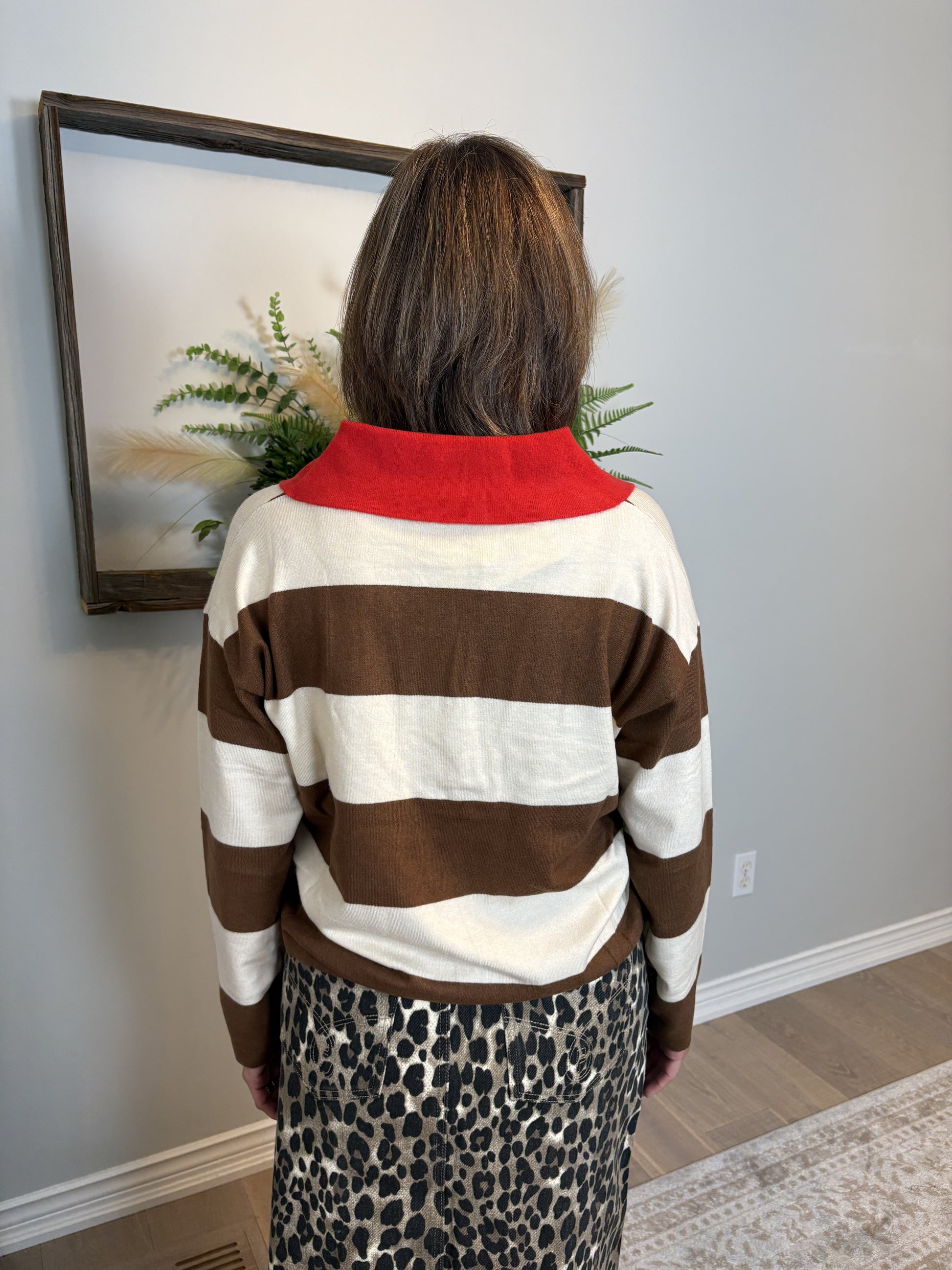 Person wearing a brown and white striped sweater with a red collar, standing in a room with a framed picture on the wall.