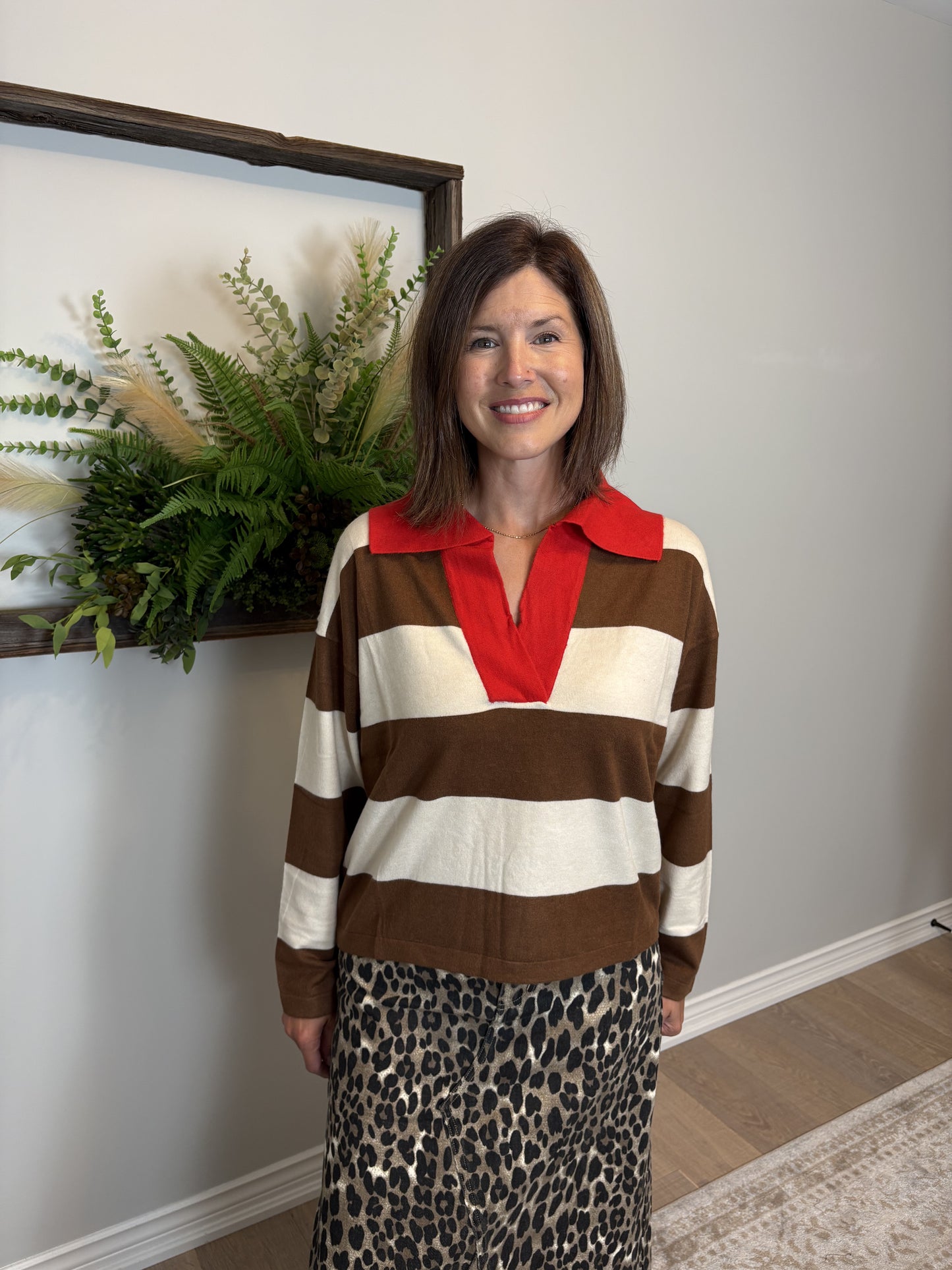 Woman wearing a striped sweater with a red collar and leopard print skirt standing in a room with a plant and framed picture on the wall.
