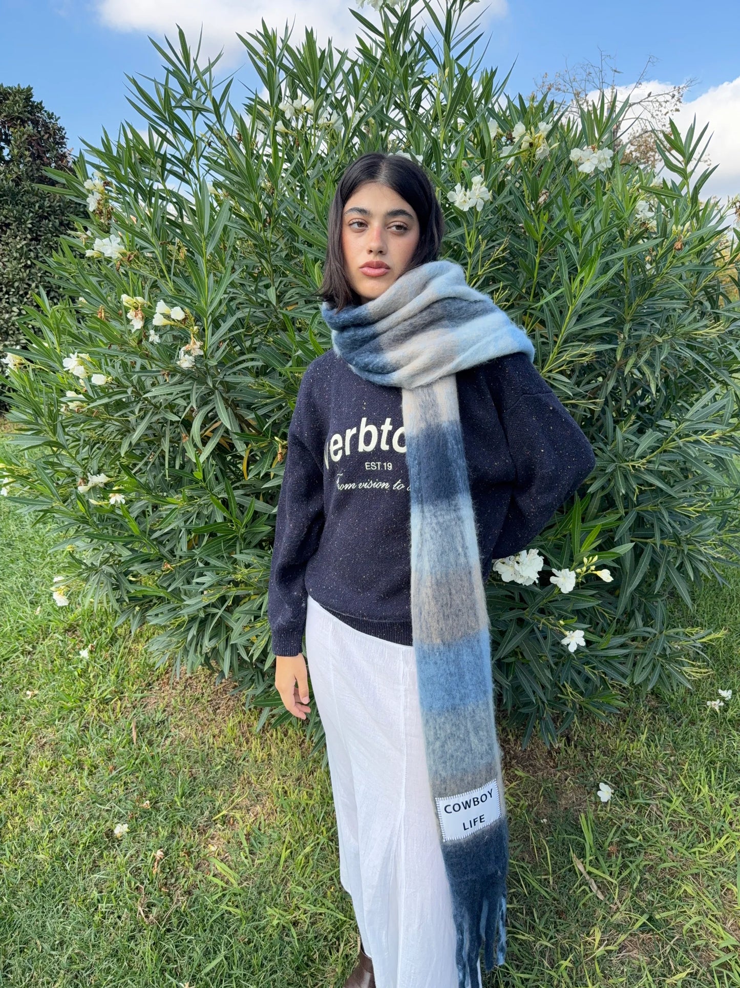 Person wearing a dark sweater with a maxi scarf in blue colours standing outdoors with greenery and flowers in the background.