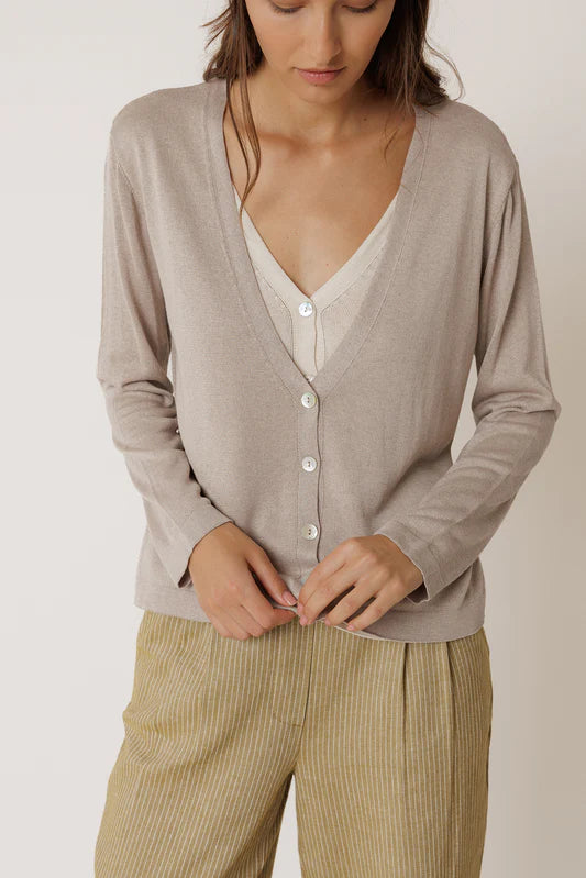 Woman wearing a beige cardigan over a white shirt with beige pants on a plain background