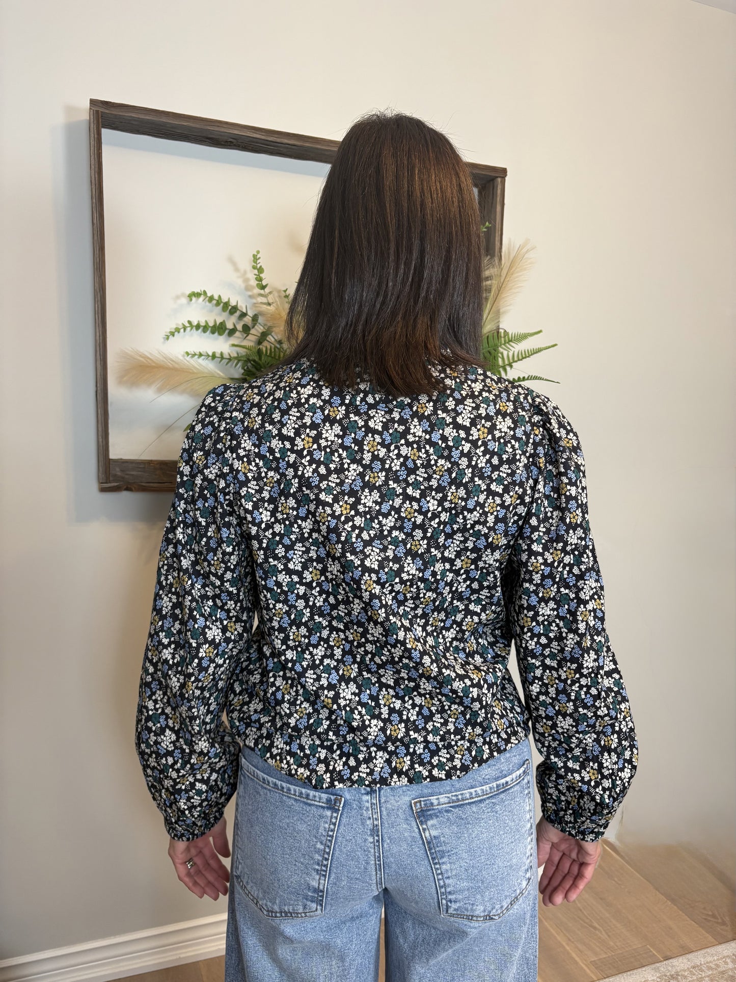Person wearing a floral blouse and jeans standing in front of a framed picture on a wall.