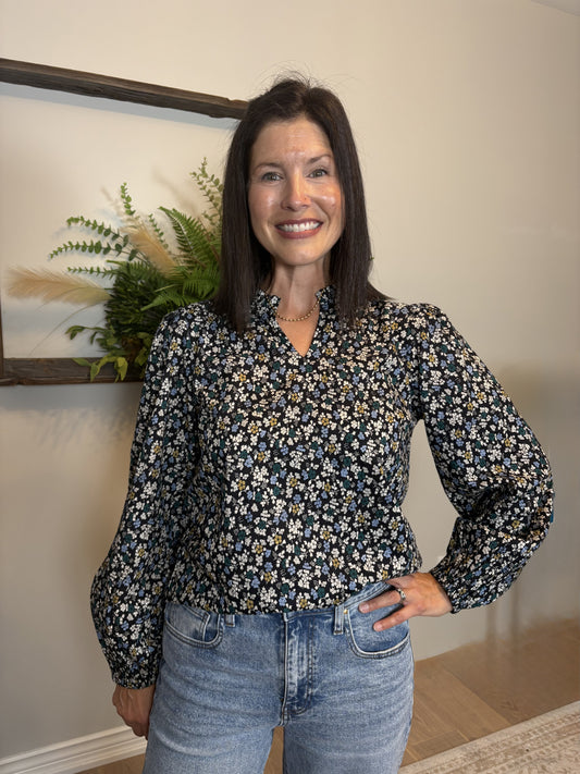 Woman wearing a floral blouse and jeans standing indoors with a plant and framed picture in the background.