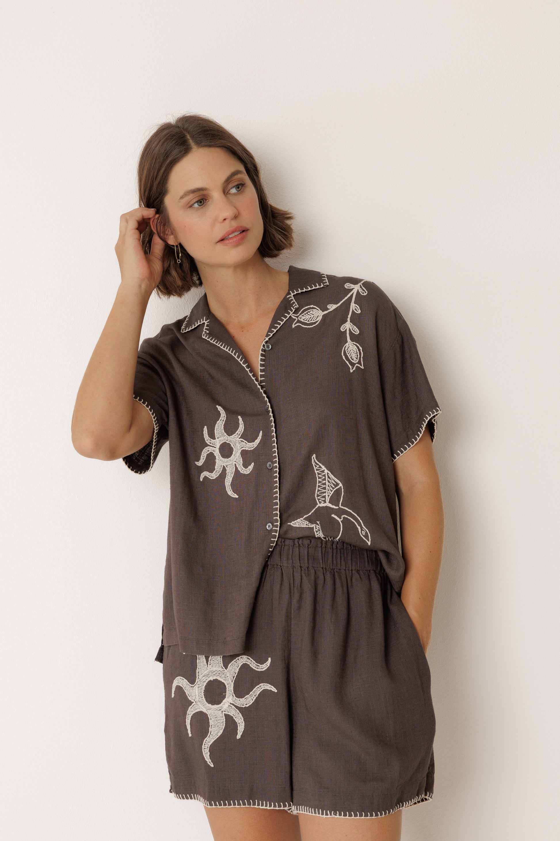 Woman wearing a brown shsorts and shirt combo with white embroidery against a white background