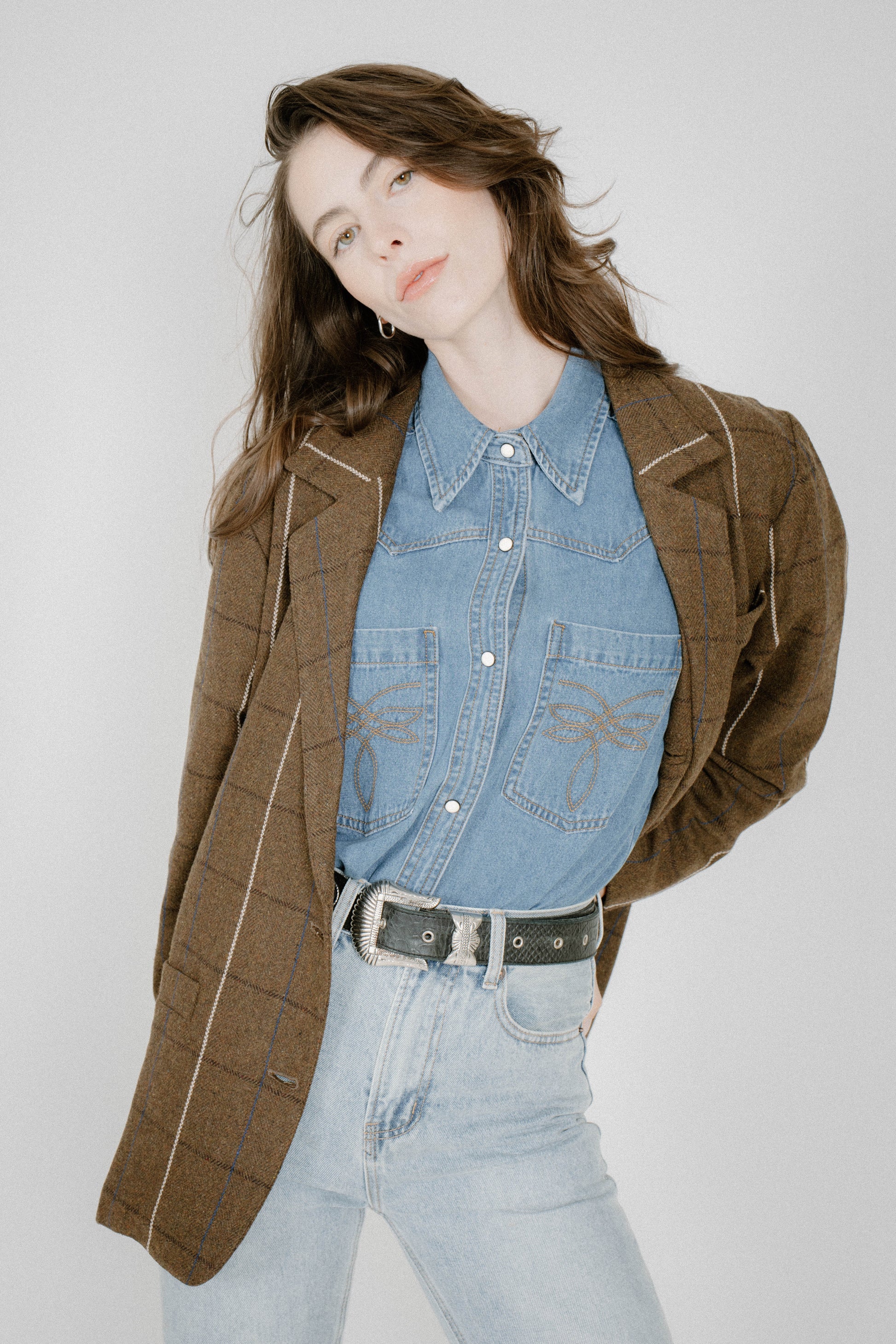 A woman modeling a brown plaid blazer over a light blue denim shirt and light-wash jeans, paired with a black belt, against a plain light gray studio background.