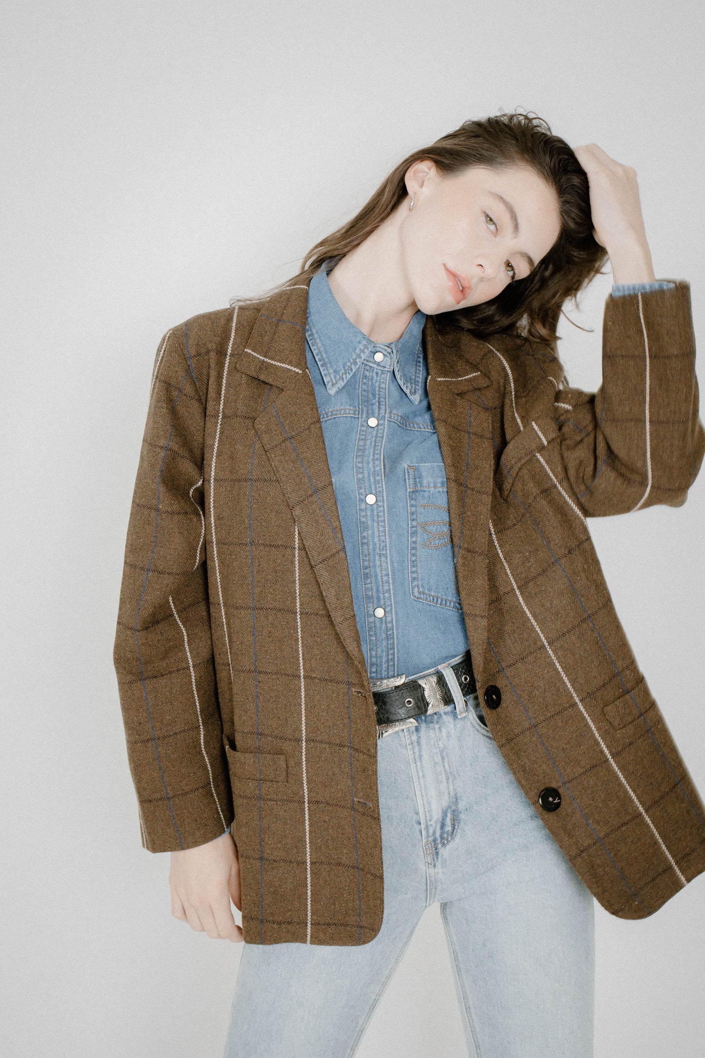 A woman wearing a brown plaid blazer with white contrast piping over a light blue denim shirt and light-wash jeans, posed against a light gray studio background.