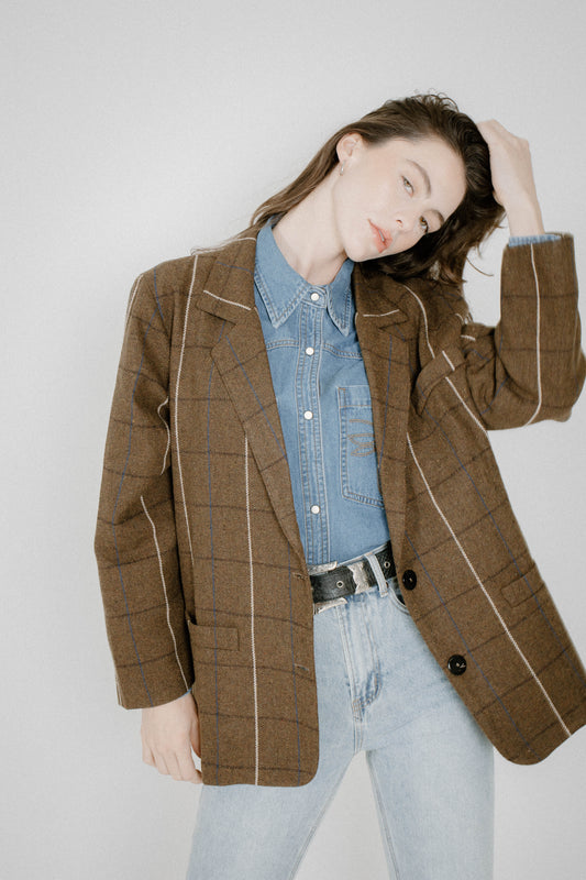 A woman wearing a brown plaid blazer with white contrast piping over a light blue denim shirt and light-wash jeans, posed against a light gray studio background.
