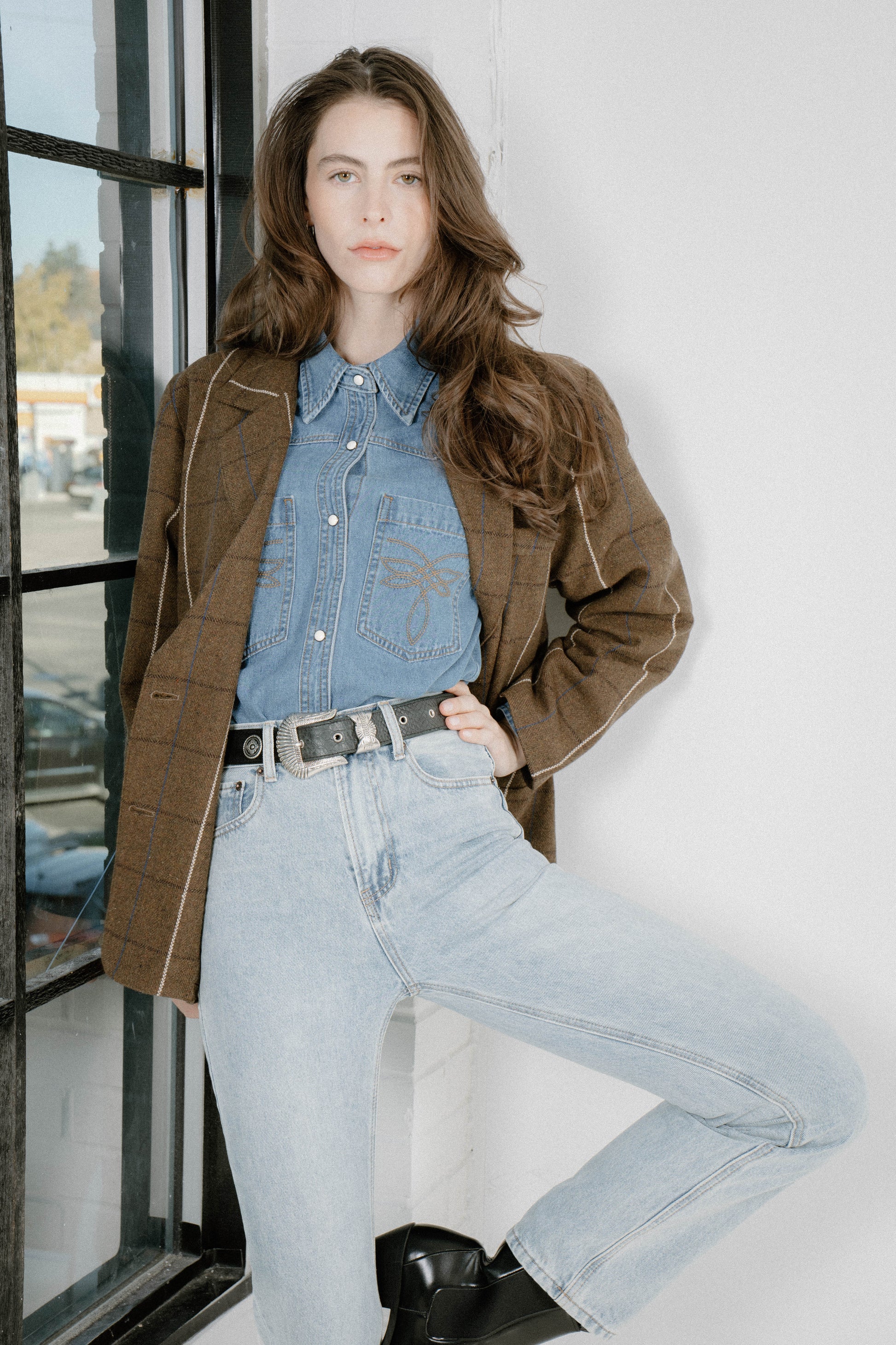 A woman models a brown plaid blazer over a light blue denim shirt and jeans, standing in a bright room next to a window.