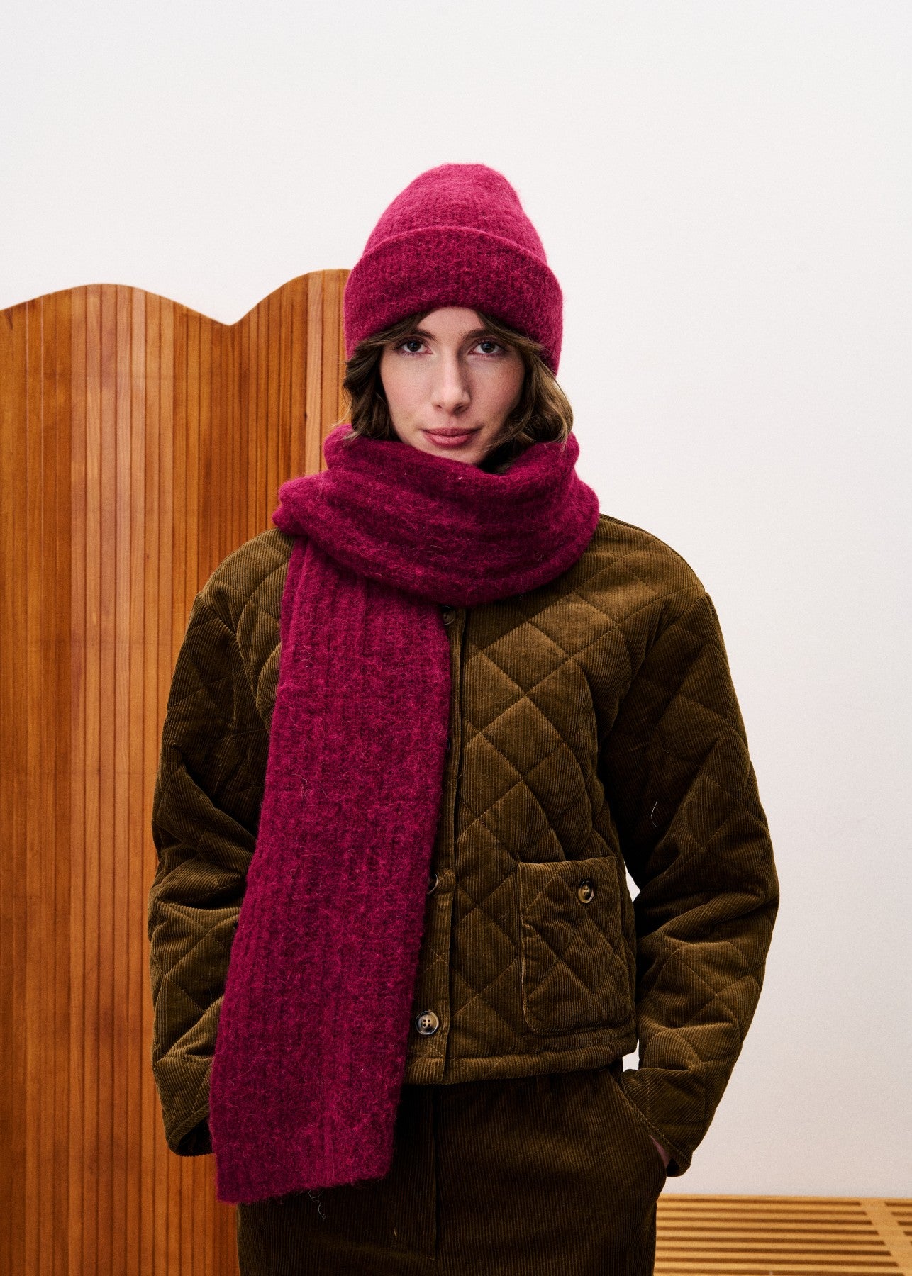 Person wearing a brown quilted jacket, pink knit hat, and pink scarf against a wooden panel background.