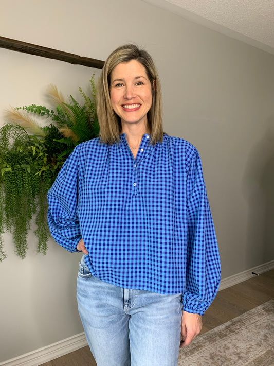 The FRNCH Noura blouse features a classic Vichy gingham pattern in blue, with long, puffed sleeves perfect for pairing with denim or a skirt for a versatile, sophisticated look. The blouse has a straight cut, round neck, making it a timeless addition to any wardrobe.