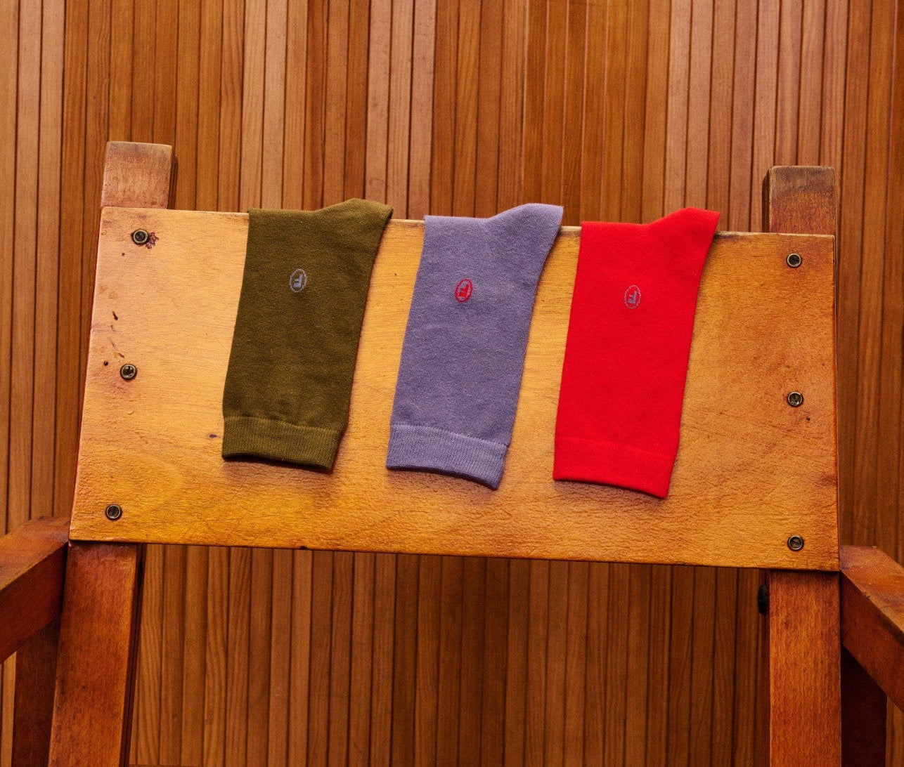 Three socks in green, blue, and red hanging on a wooden bench against a wooden paneled wall.