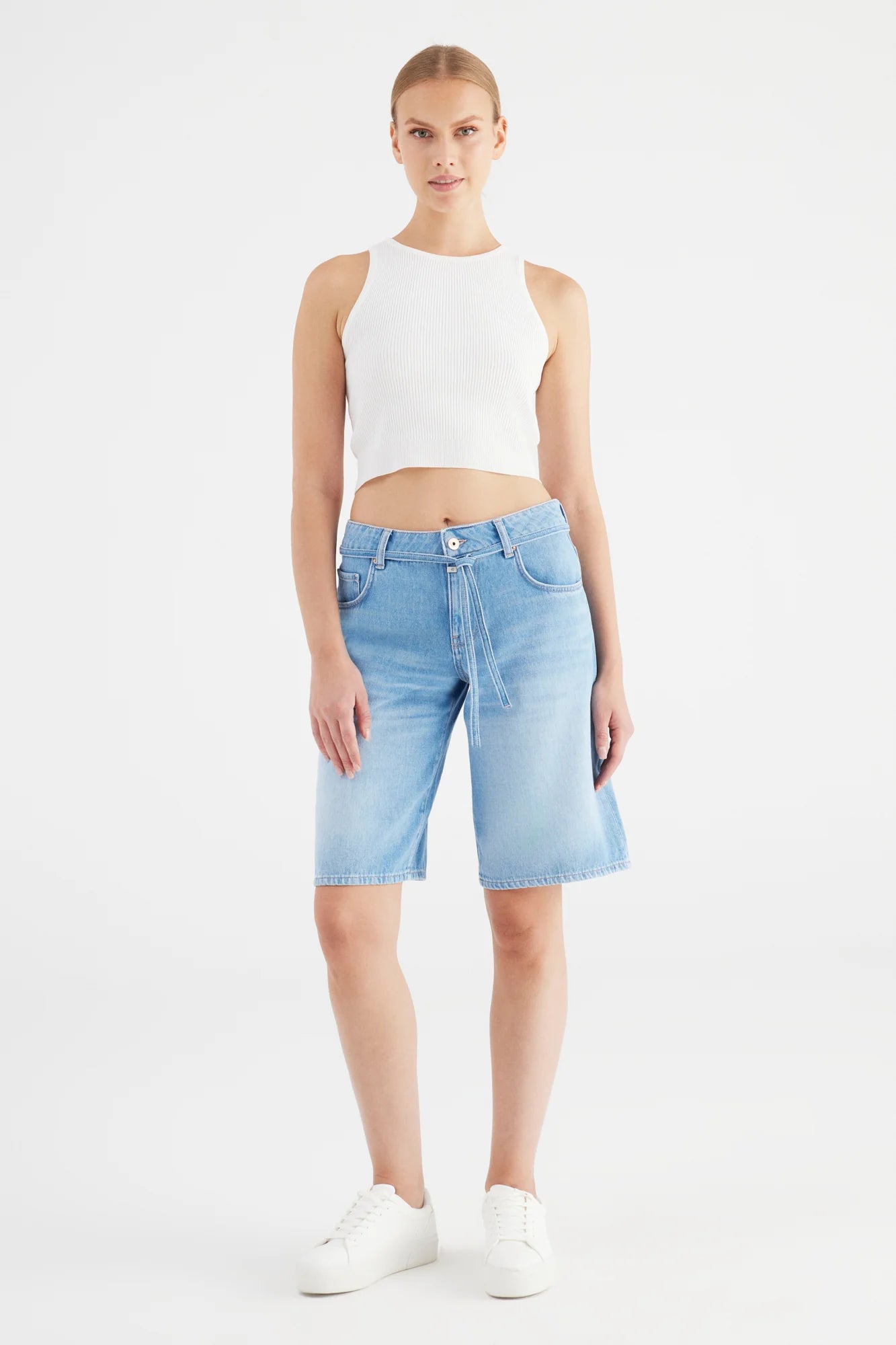 Woman wearing a white sleeveless top and light blue denim shorts on a white background