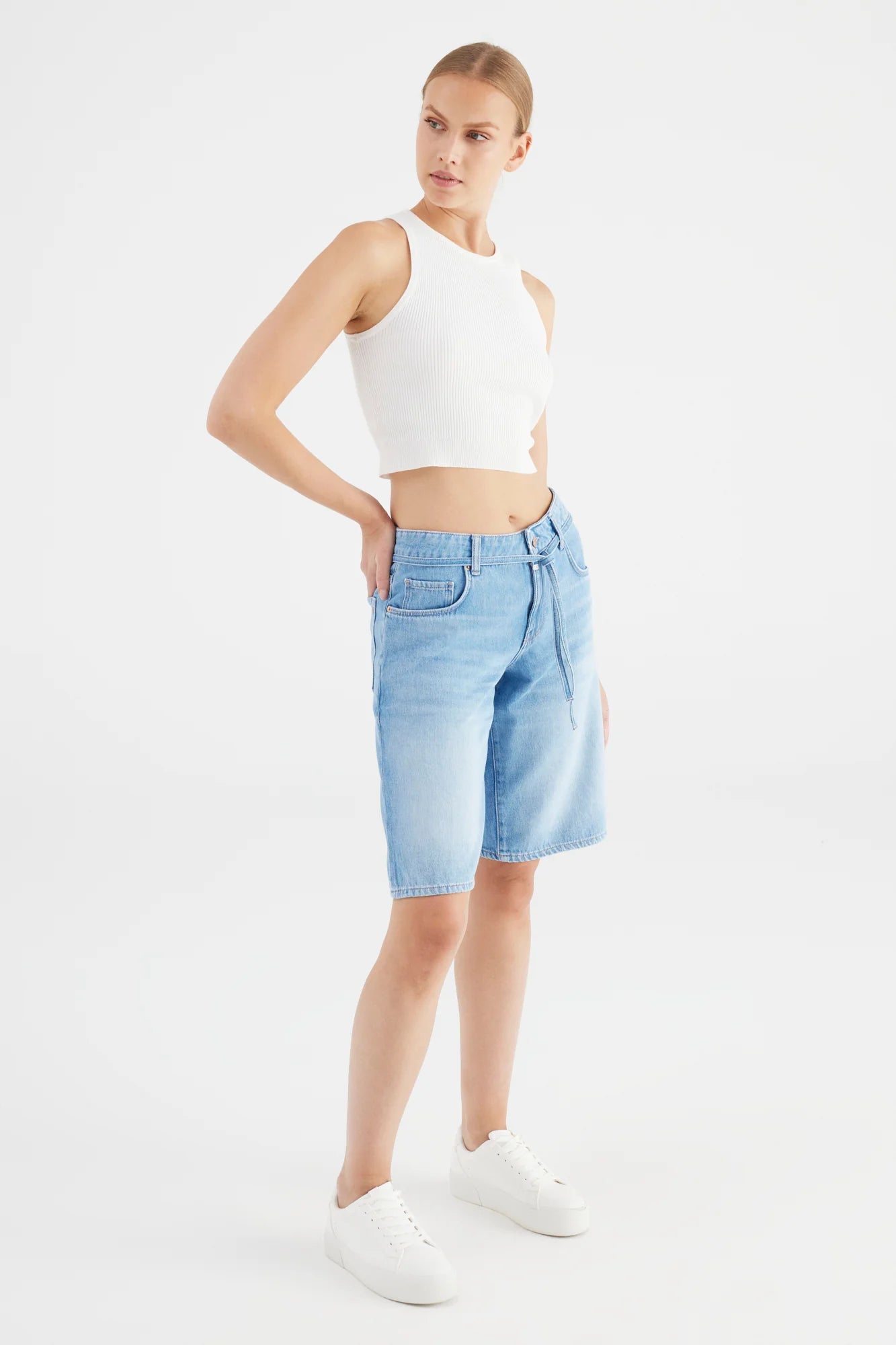 Woman wearing a white sleeveless top and light blue denim shorts on a white background