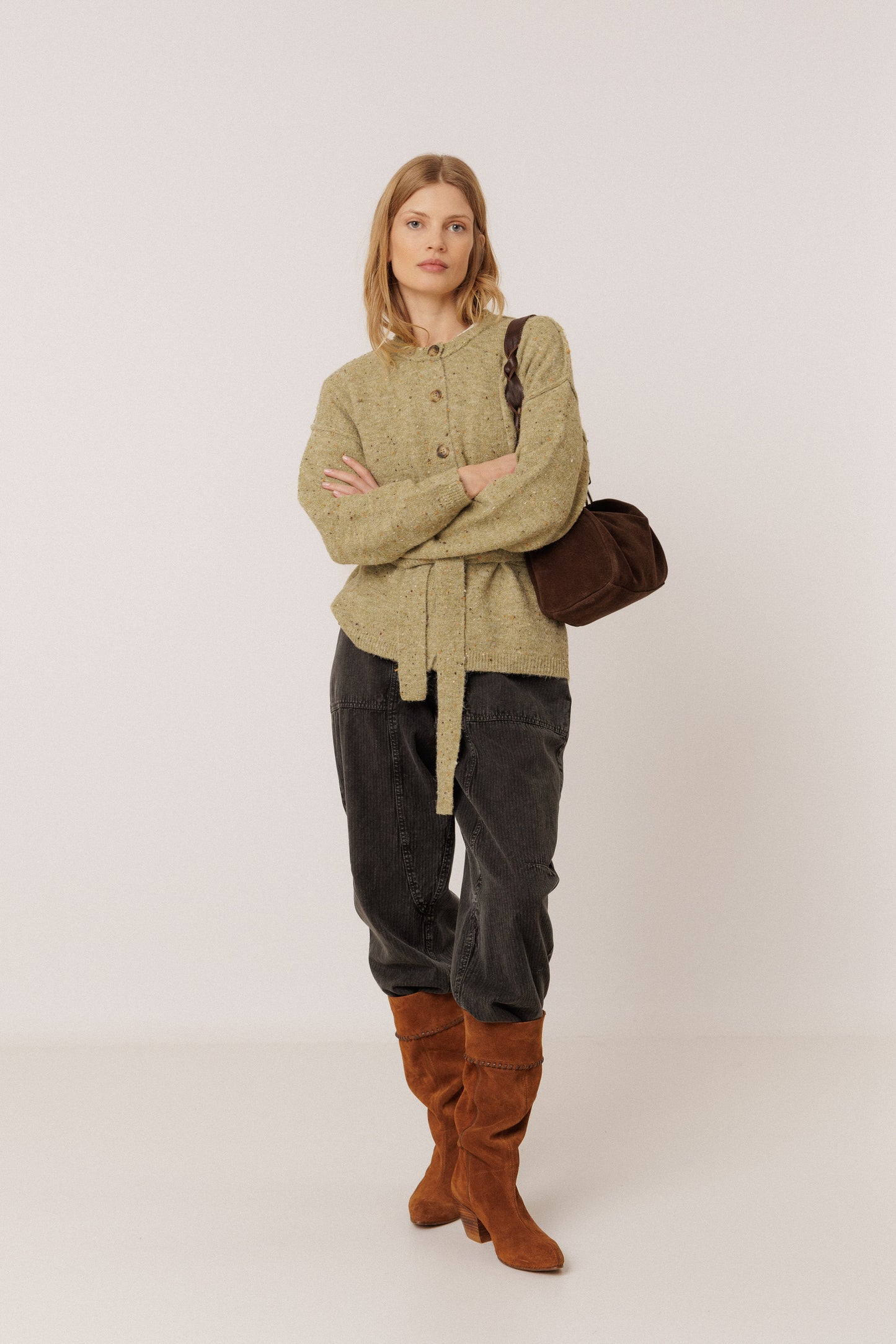 A model stands against a light gray studio background wearing a textured olive green cardigan, dark gray pants, brown suede boots, and carrying a dark brown shoulder bag.