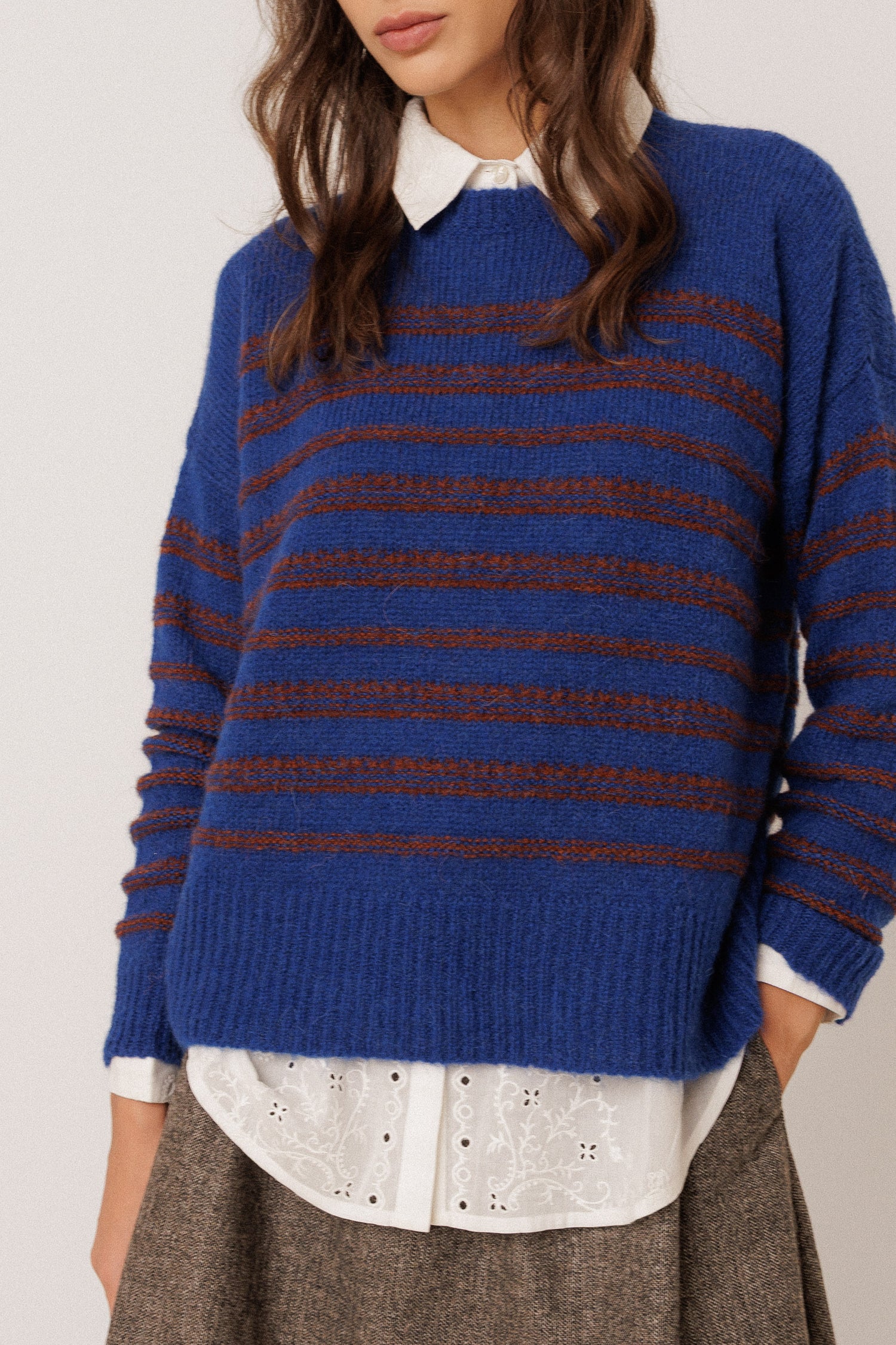 A woman wearing a blue knit sweater with horizontal brown stripes, layered over a white collared shirt and brown skirt, against a plain light background.