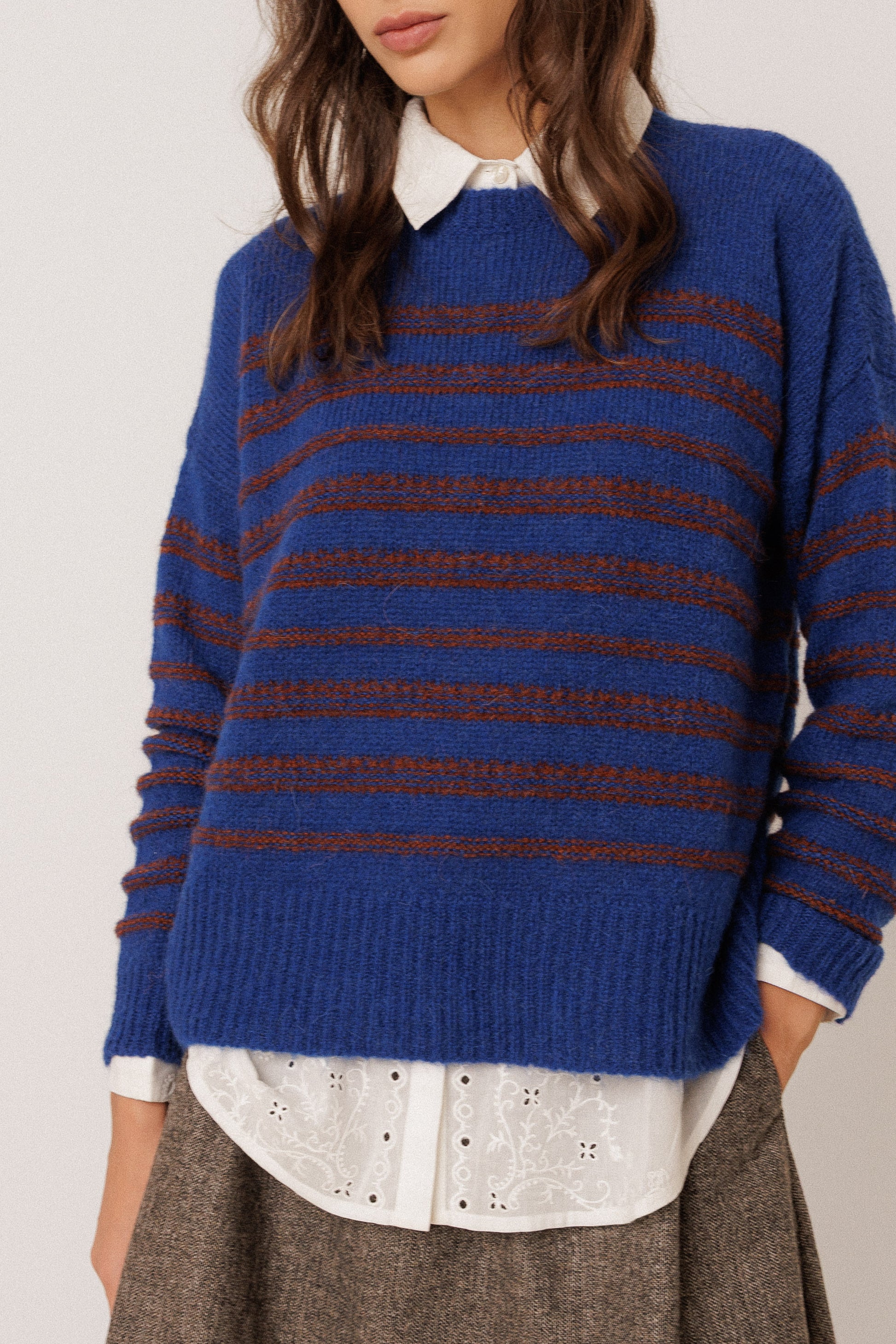 A woman wearing a blue knit sweater with horizontal brown stripes, layered over a white collared shirt and brown skirt, against a plain light background.