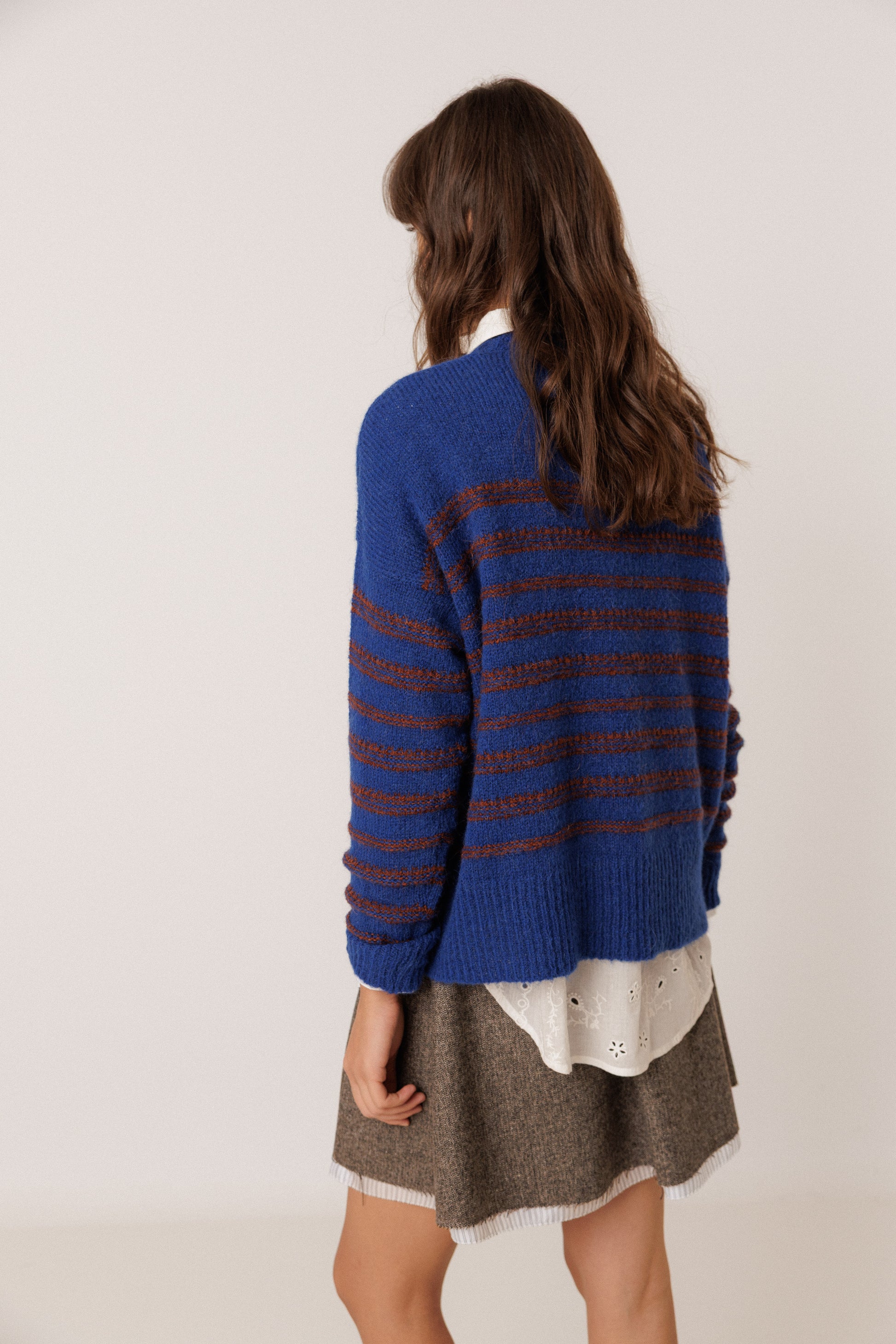 A woman shown from the back wearing a blue knit sweater with horizontal dark stripes, a white undershirt, and a brown skirt against a plain light background.