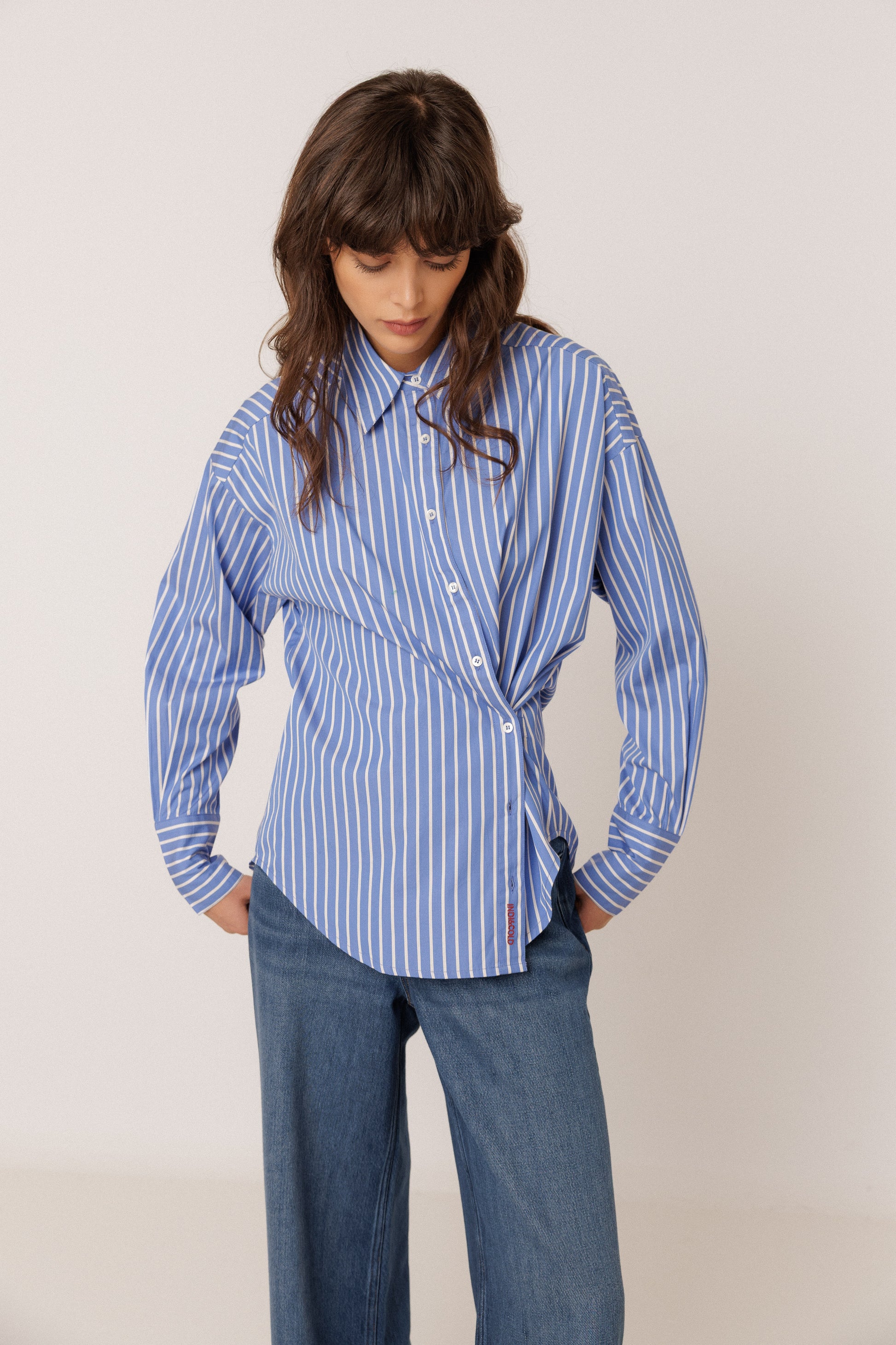A woman wearing a blue and white vertically striped button-up shirt with a twisted waist detail, paired with blue jeans, standing against a plain light beige background.