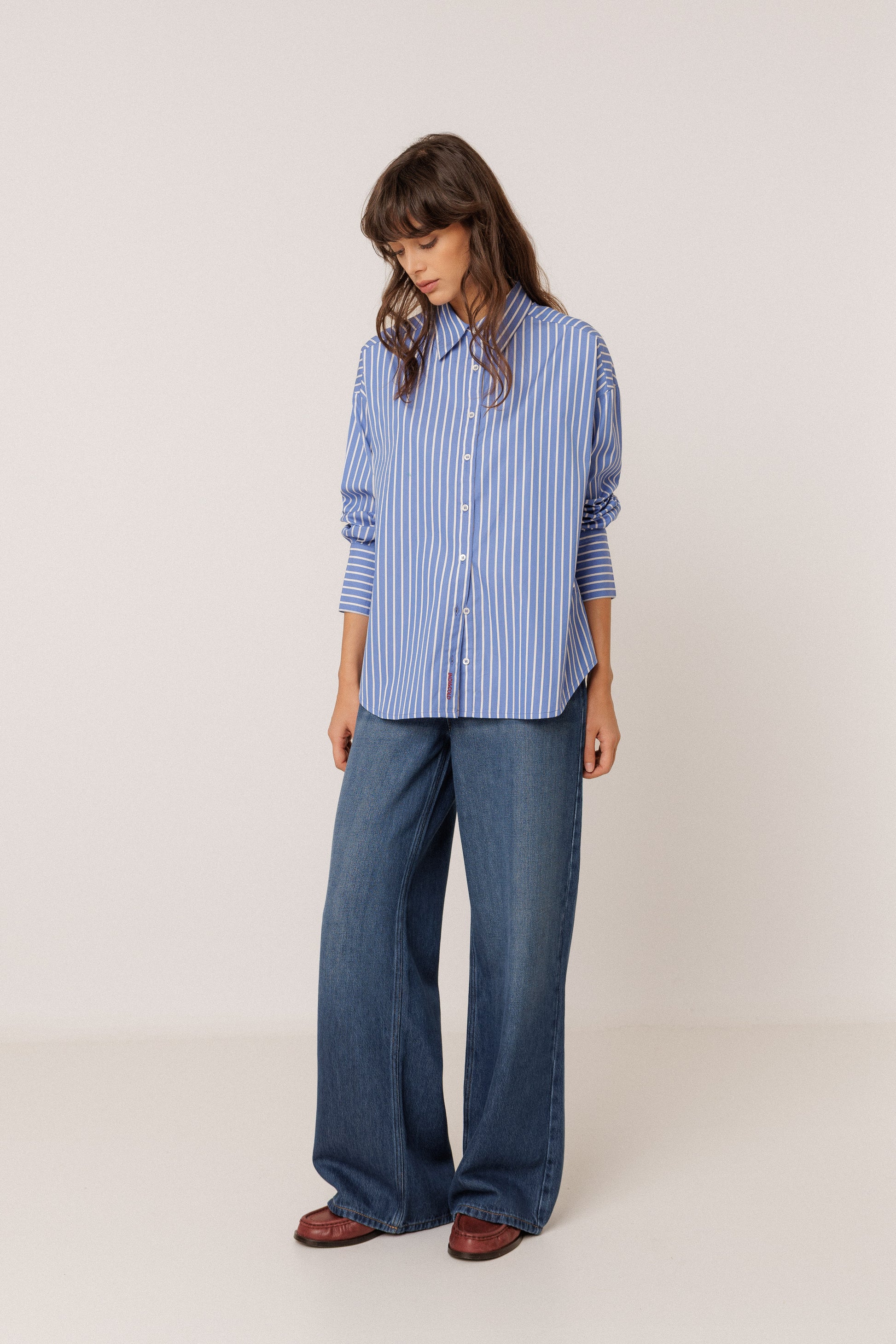 A woman modeling a blue and white vertically striped button-down shirt with wide-leg blue jeans and brown shoes, standing against a plain light beige studio background.