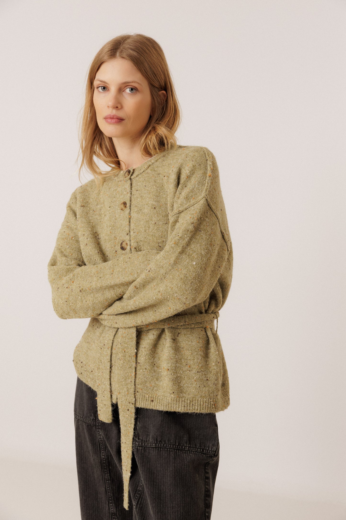 A woman modeling a textured olive green knit cardigan with a buttoned front, drawstring waist, and dark charcoal pants against a plain light background.