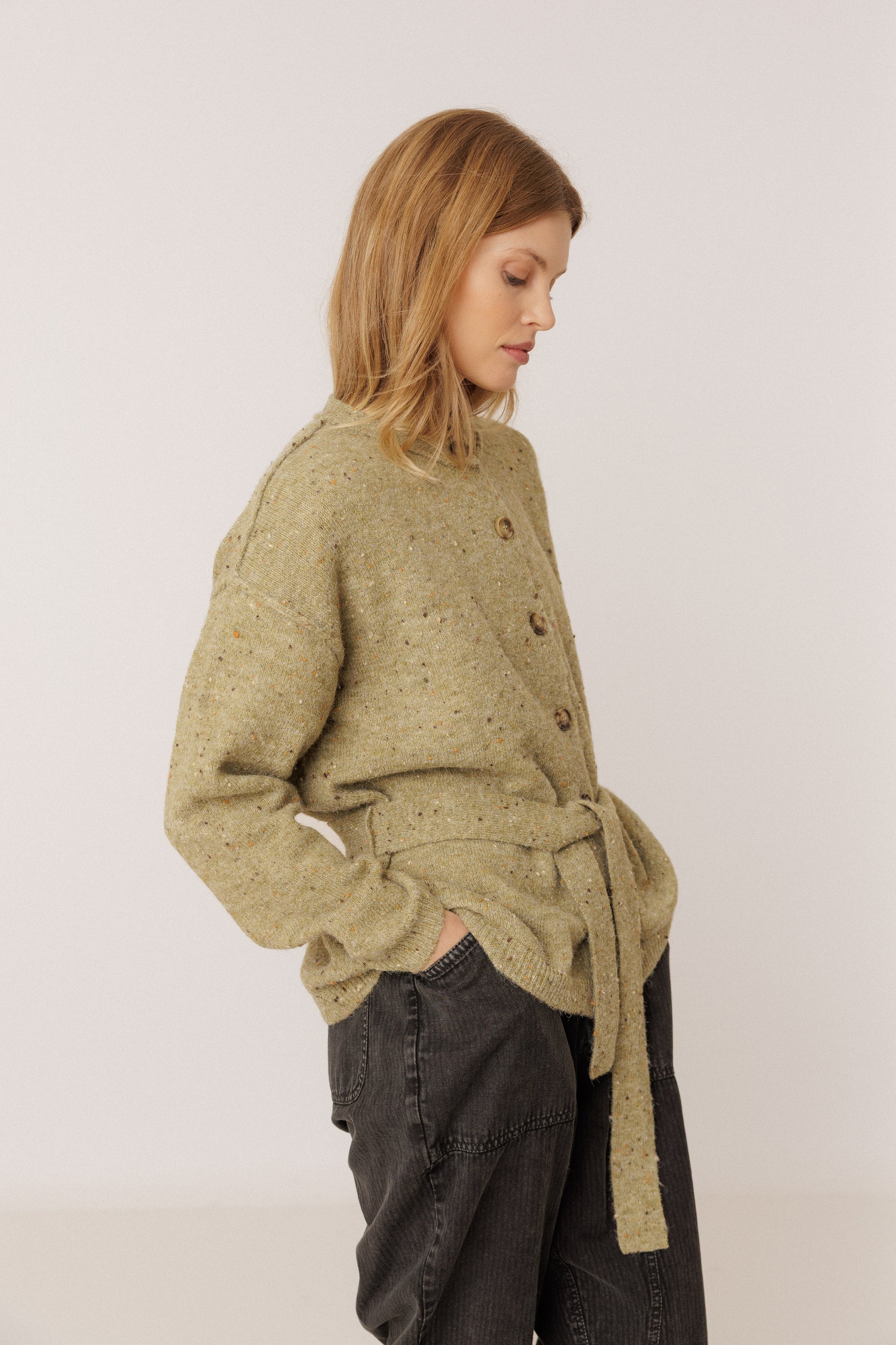 A woman wearing a textured olive green knit cardigan with a tie waist and dark gray jeans, photographed against a plain light gray studio background.