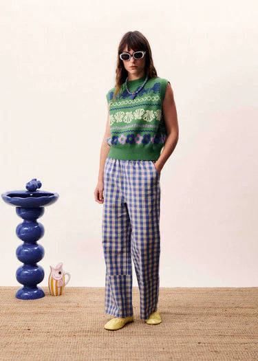 Person wearing a green patterned sleeveless top and blue checkered pants standing in a room with a decorative blue vase and pink wall.