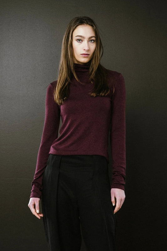 Person wearing a maroon turtleneck sweater and black pants against a dark background