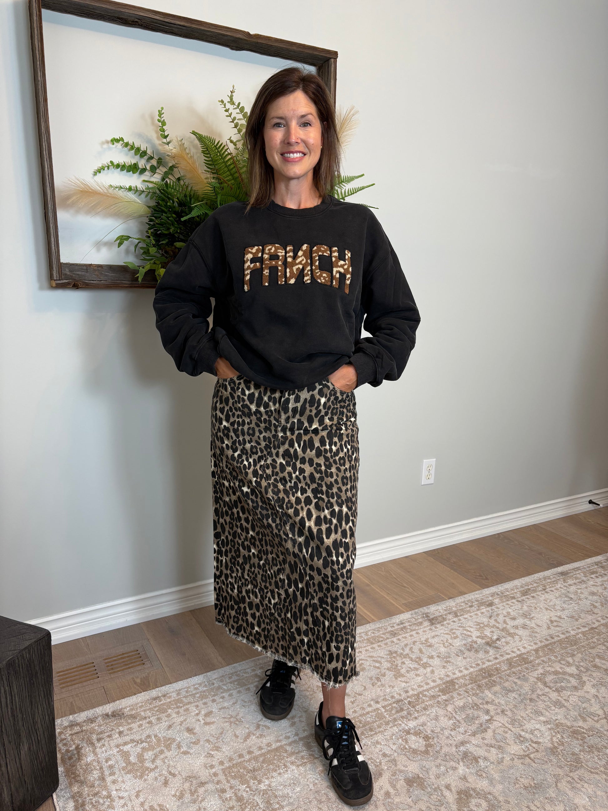 Woman wearing a black sweatshirt with 'FRNCH' and a leopard print skirt indoors.