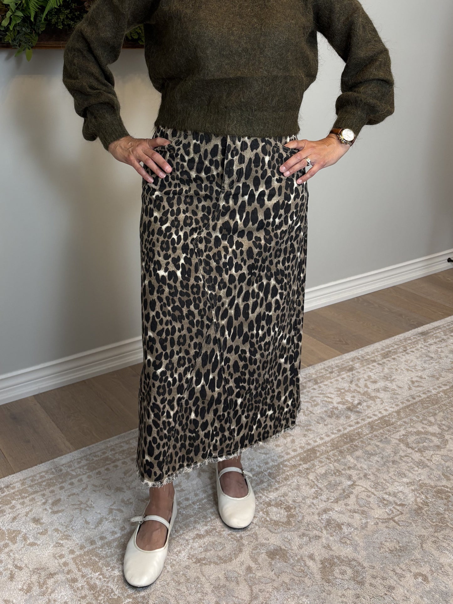 Woman in a green sweater and leopard print skirt standing in a room with a painting and plant on the wall.