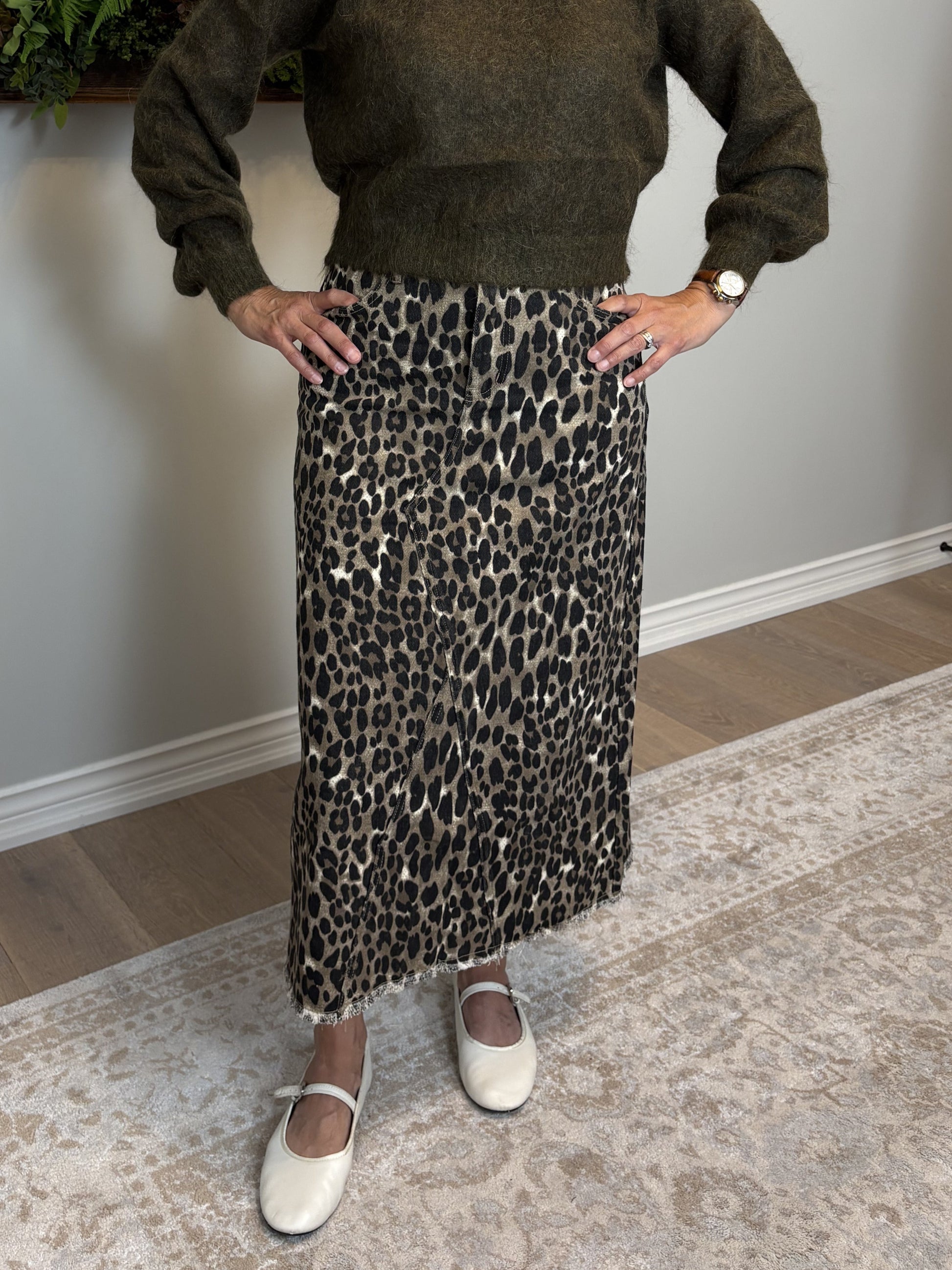 Woman in a green sweater and leopard print skirt standing in a room with a painting and plant on the wall.