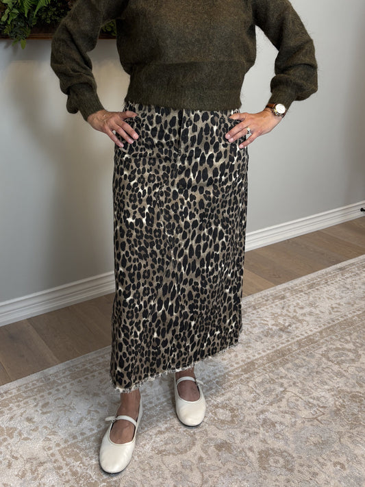 Woman in a green sweater and leopard print skirt standing in a room with a painting and plant on the wall.
