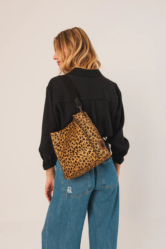 Person wearing a black jacket and blue jeans with a leopard print bag on a plain background
