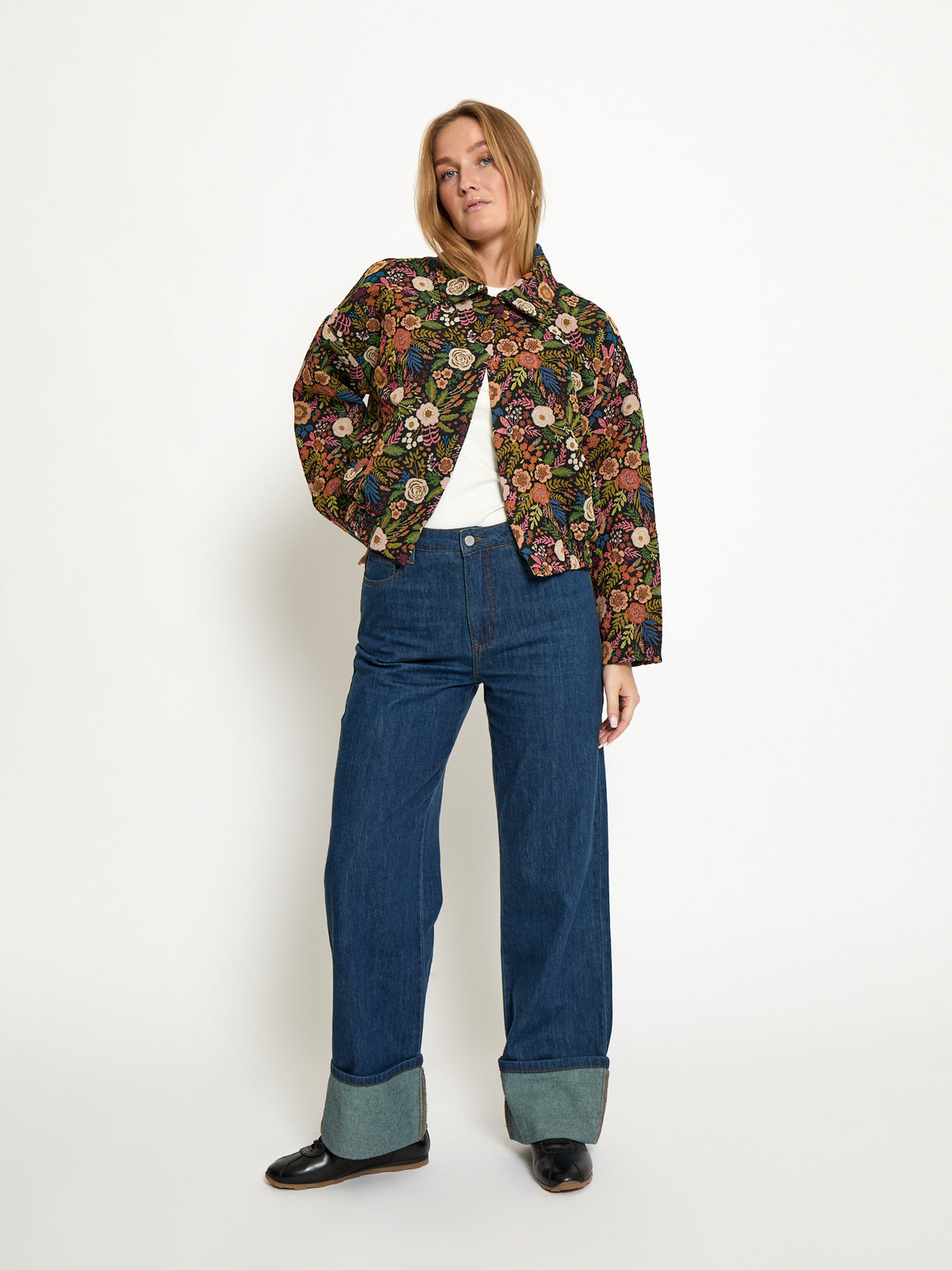 A woman modeling a cropped, long-sleeve button-up jacket with an intricate multicolor floral and paisley pattern, paired with blue jeans featuring rolled cuffs and black shoes, against a plain white studio background.