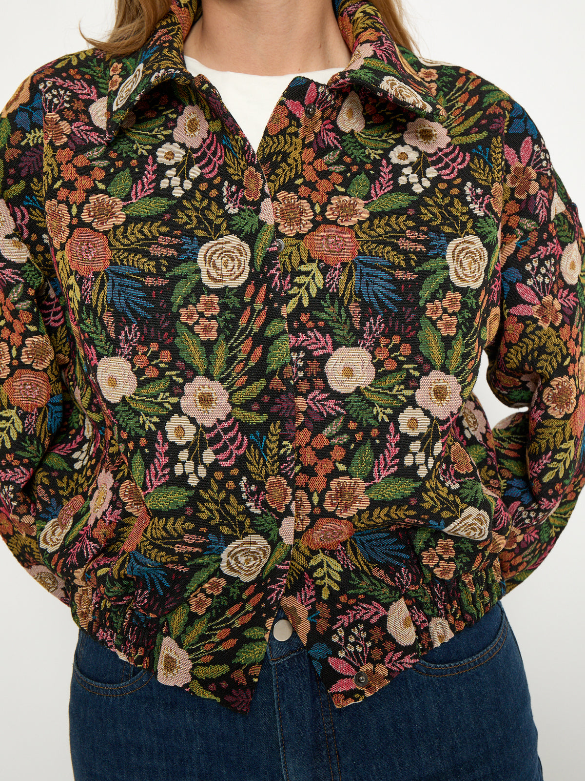 A close-up of a person wearing a long-sleeve button-up shirt with an intricate, dense floral and botanical pattern in green, pink, orange, and cream, paired with blue jeans.