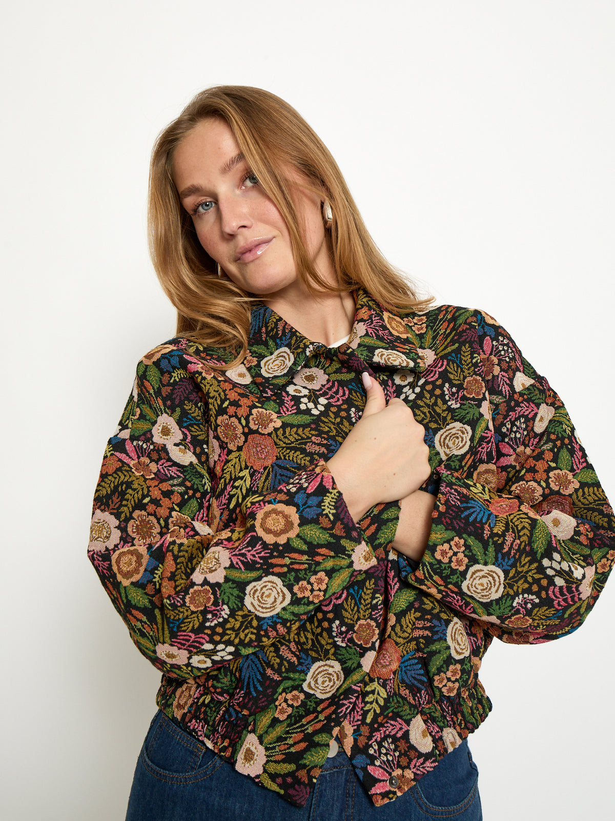 A woman wearing a long-sleeve button-up blouse with a dense multicolor floral and botanical print, paired with blue jeans, photographed against a plain white studio background.