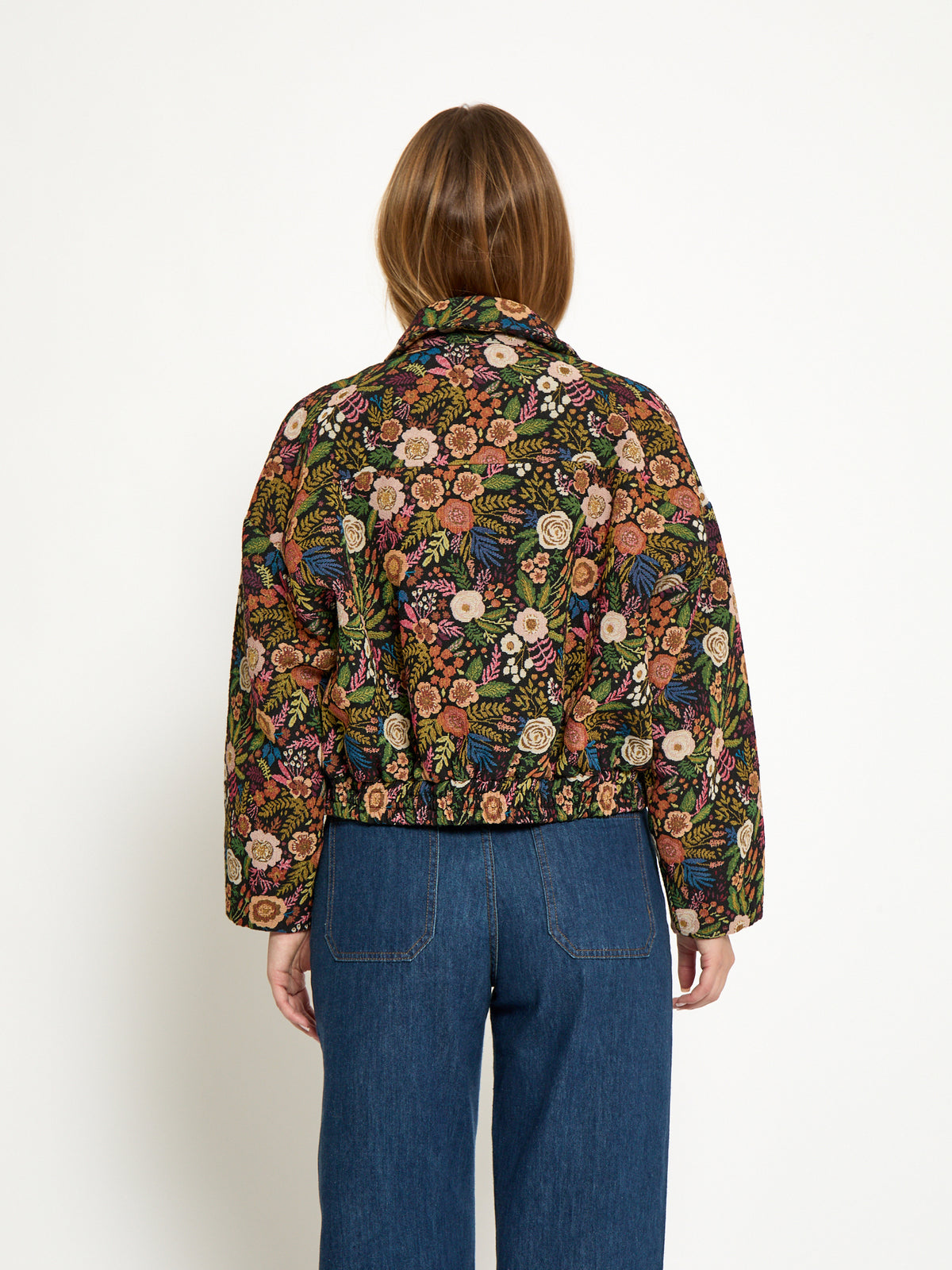 A model viewed from the back wearing a cropped, long-sleeve button-up shirt with an intricate multicolor floral and botanical pattern, paired with blue jeans, against a plain white background.