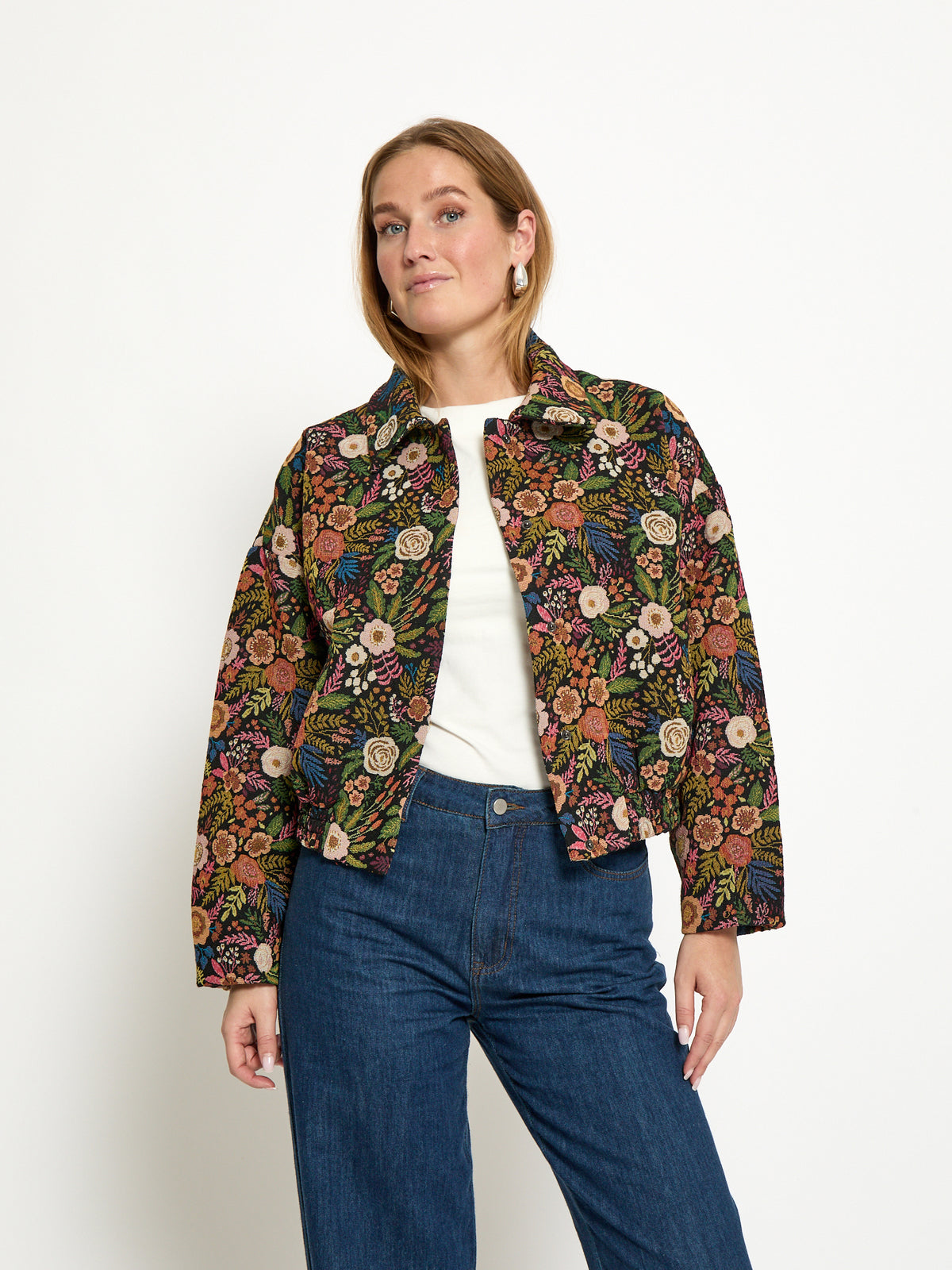 A woman modeling a cropped, long-sleeve button-up jacket with an intricate multicolor floral and paisley pattern on a dark background, styled with a white top and blue jeans against a plain white studio background.