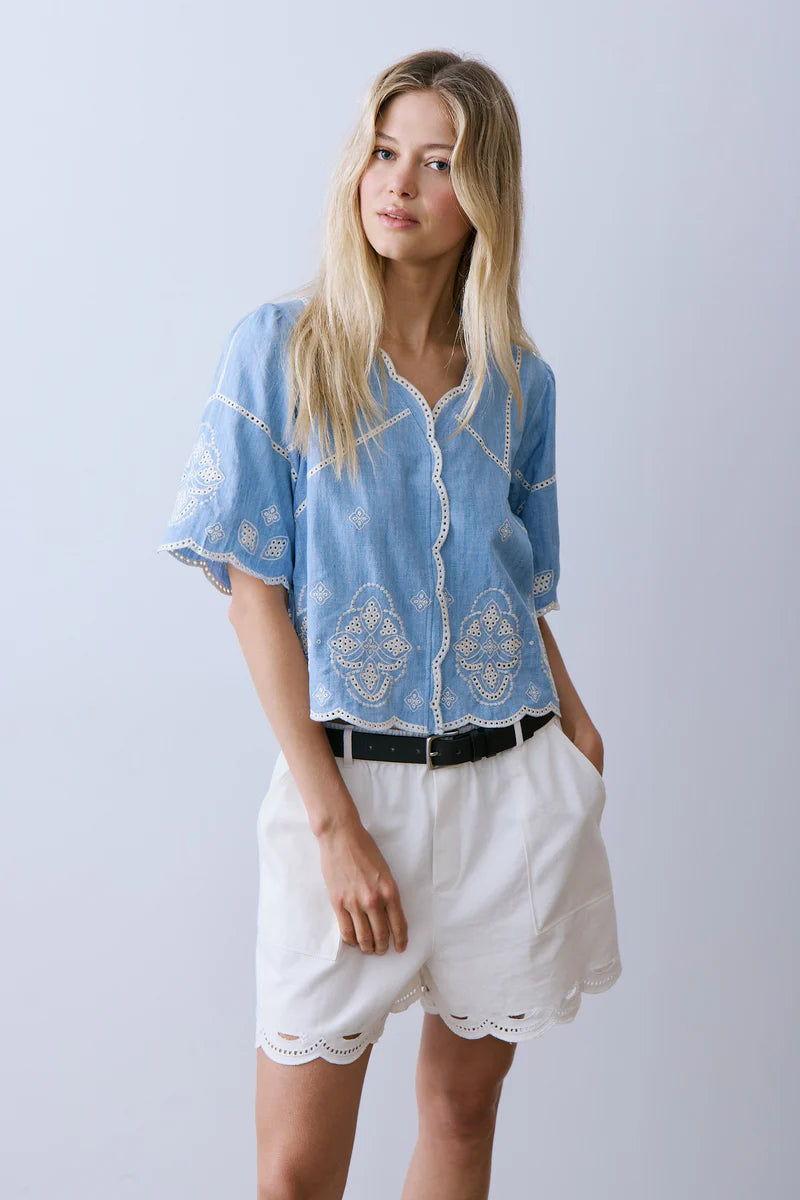 Woman wearing a blue embroidered top and white shorts on a light gray background