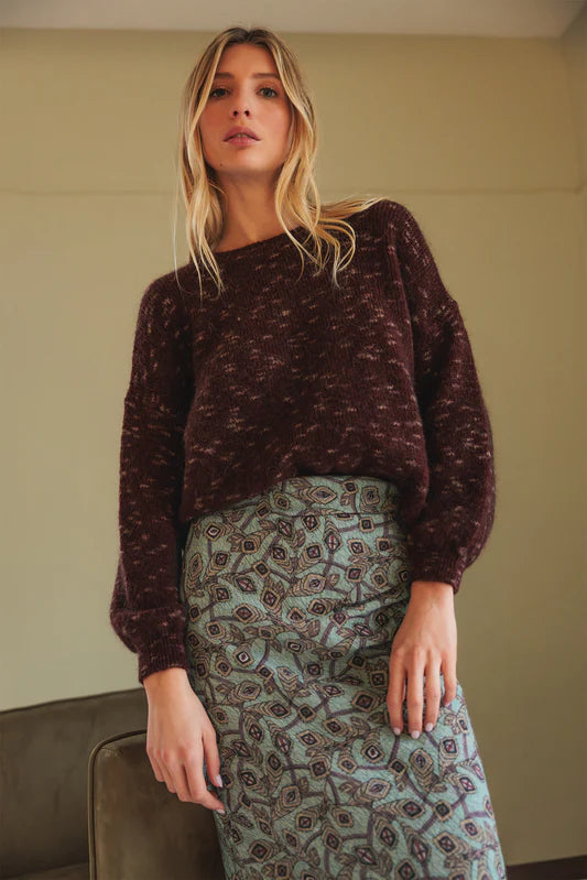 Woman wearing a brown sweater and patterned skirt indoors.