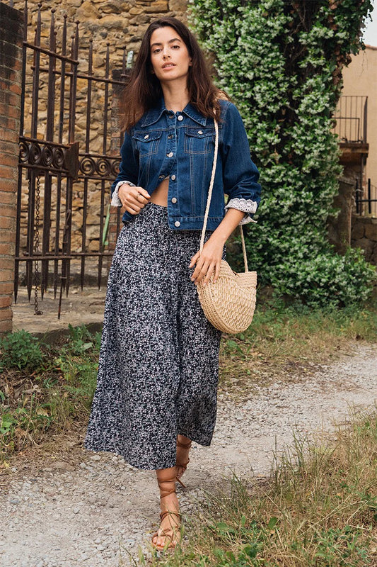 Woman in denim jacket and floral skirt holding a woven bag outdoors.