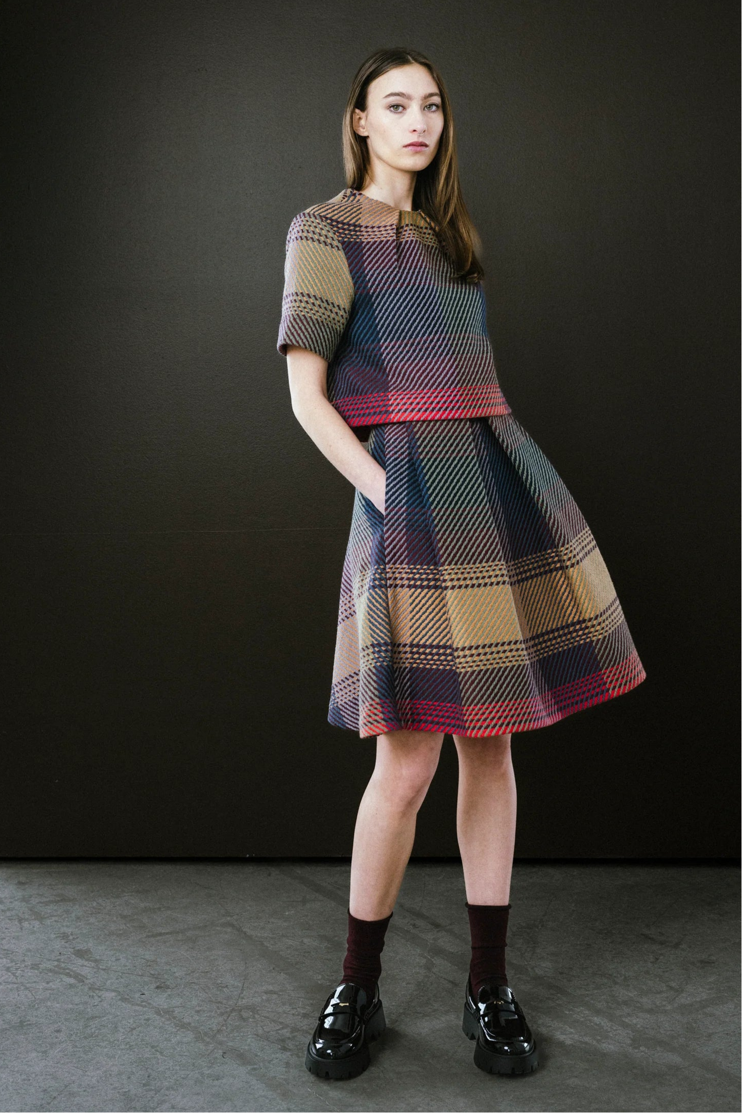 Woman wearing a plaid dress standing against a black wall.