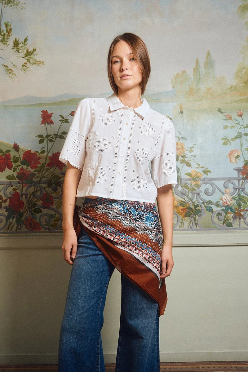 Woman wearing a white blouse and blue jeans with a patterned scarf, standing in front of a decorative wall.