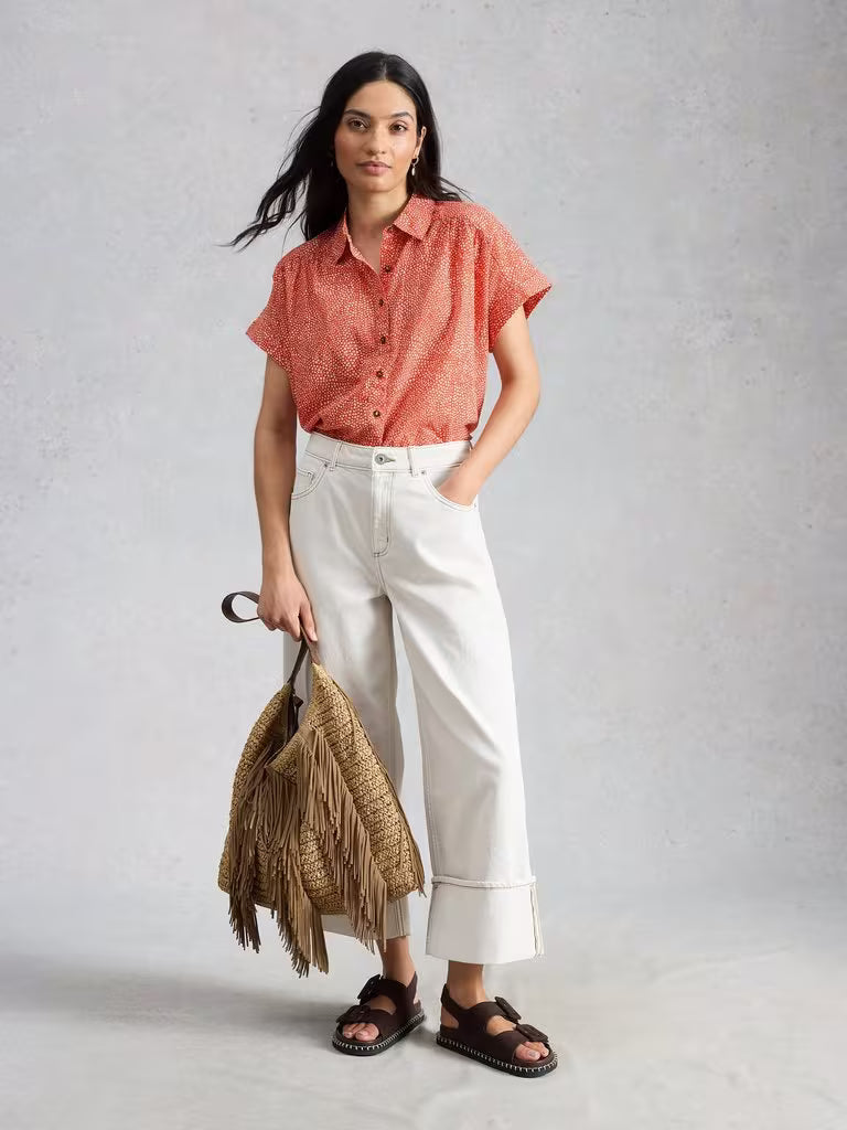 Woman wearing a red patterned shirt and white pants, holding a woven bag against a gray background