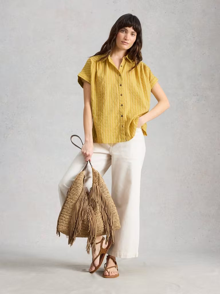 Woman wearing a yellow shirt and white pants, holding a straw bag against a plain background
