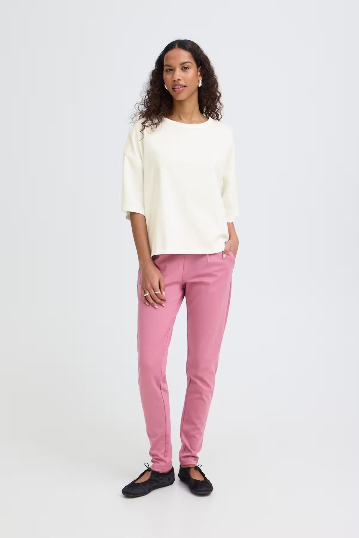 A woman modeling a cream-colored short-sleeve top and pink pants against a light gray studio background.