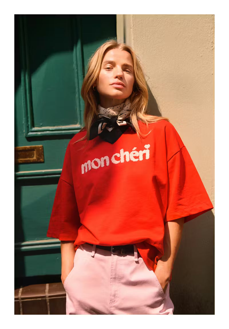 Person wearing a red 'mon chéri' shirt standing against a green door and beige wall.
