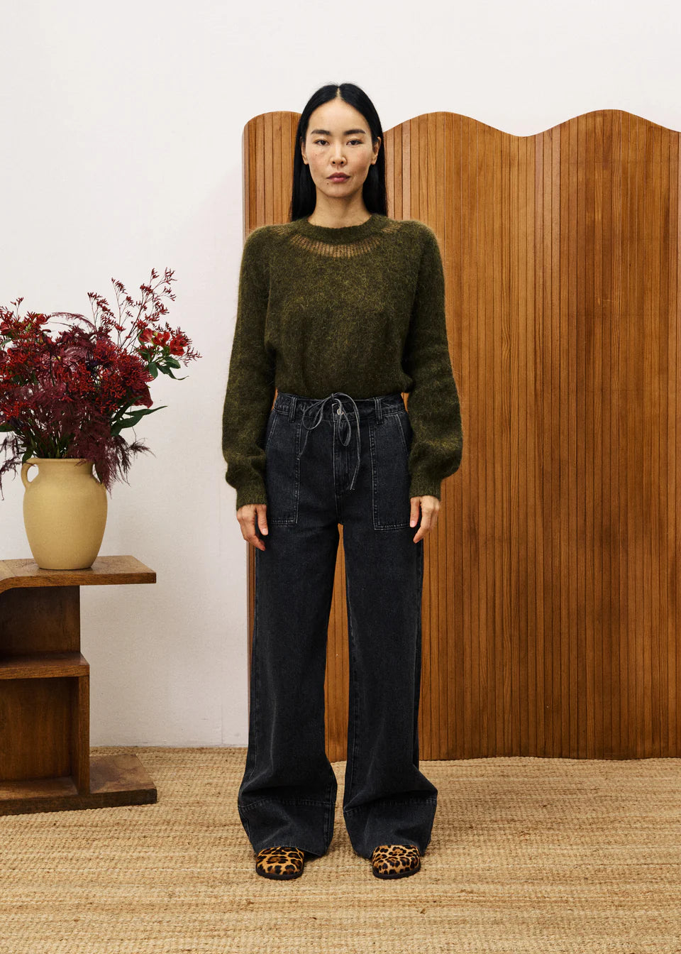 Woman wearing a green sweater and black pants standing in front of a wooden screen with a vase of flowers on a table.