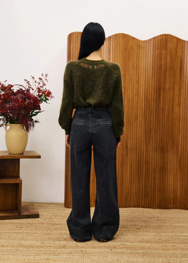Person wearing a green sweater and black jeans standing in front of a wooden screen with a vase of flowers on a table.